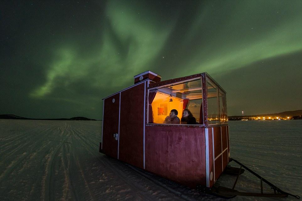 Пара в освещённом домике Lake Inari со снегоходом под зелёным северным сиянием