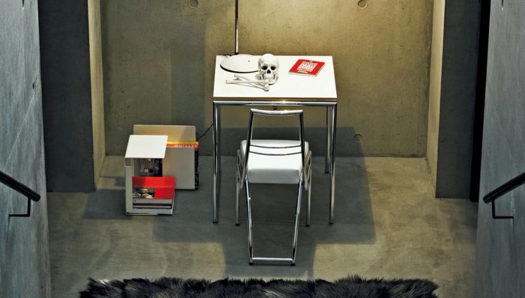 A writing desk with chromed legs and a chair with a skeleton backrest, a skull and books in a concrete alcove
