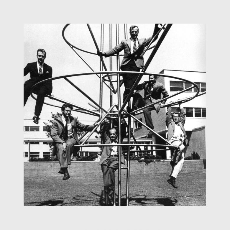 Six men in suits seated on a metal spiral sculpture in the courtyard of the TAC office