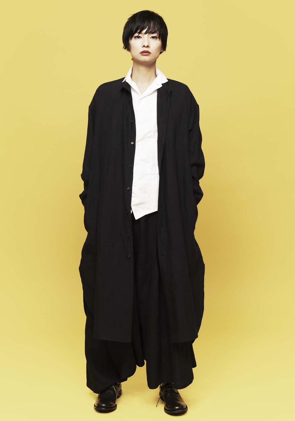 A model in a long black shirt-coat worn over a white blouse, against a yellow background