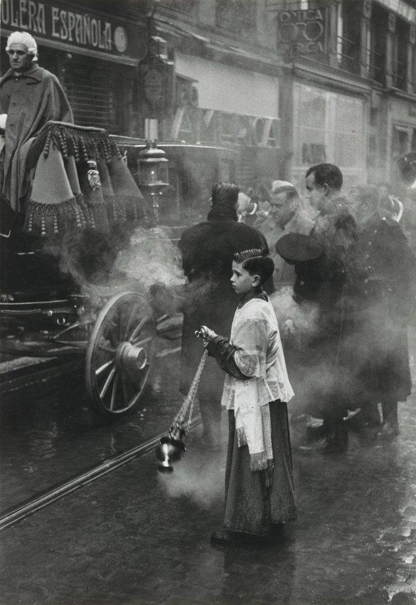 An altar boy carrying a censer stands beside a funeral carriage amid billowing smoke on a city street