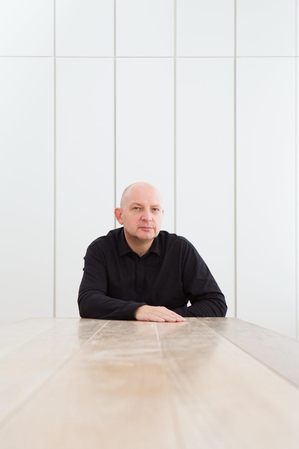 Portrait of a bald man in black seated at a wooden table against a white wall — architect Claudio Silvestrin