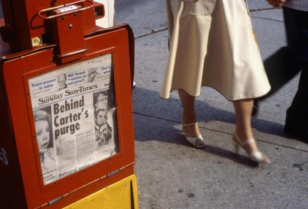 Colour photograph: a red newspaper stand with a headline, and the legs of a woman in light sandals