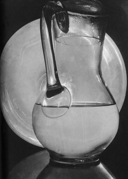 A still life with a glass pitcher of water against a pale plate