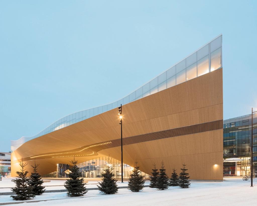 The curved wooden façade of Oodi library in Helsinki in winter, with snow-covered fir trees in front of the entrance