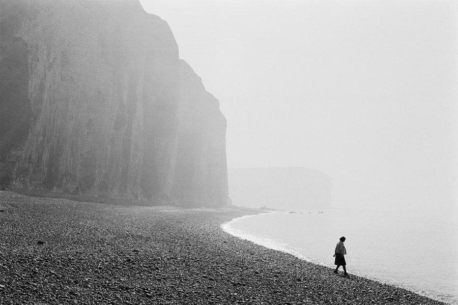The solitary figure of a woman walking along a pebble beach at the foot of tall cliffs in the mist
