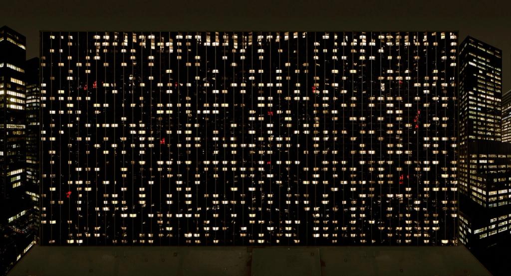 The night-time façade of a skyscraper, its grid of lit windows set between dark buildings