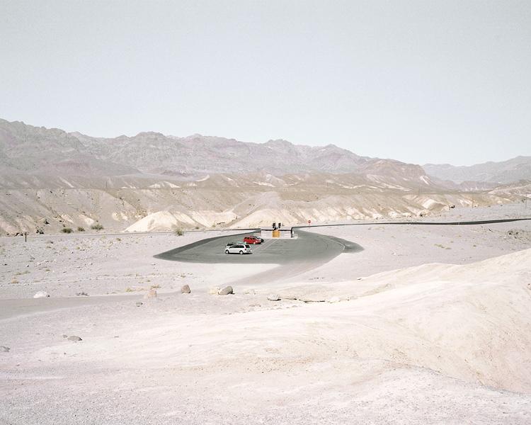 Two cars on a circular lot in the middle of a pale, stony desert with mountains behind