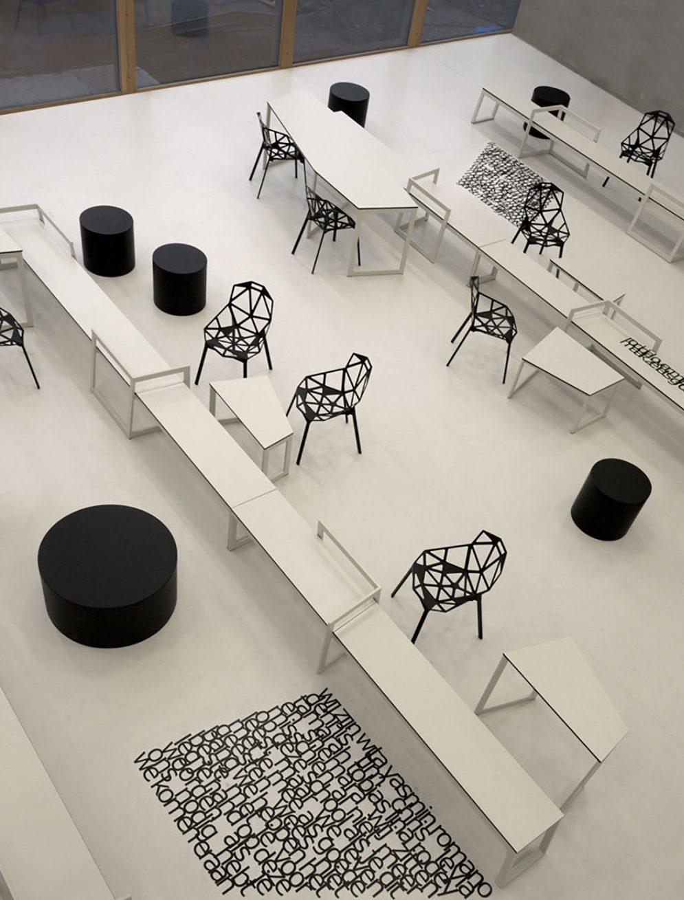 Panta Rhei School: a white interior with black openwork chairs, tables and letters on the floor, overhead view