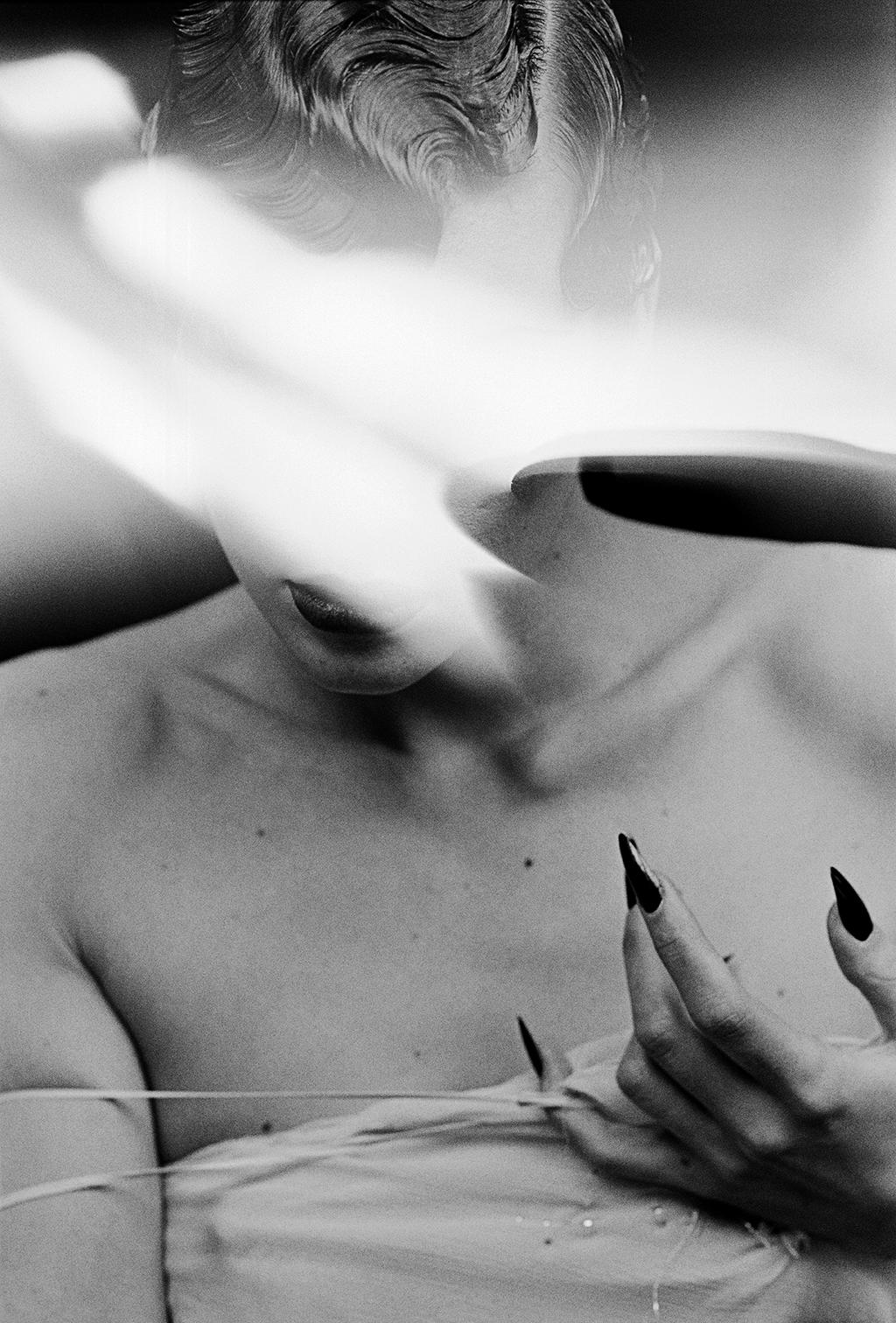 A black-and-white frame of a model in a light-coloured outfit, a hand with long black nails resting at her chest
