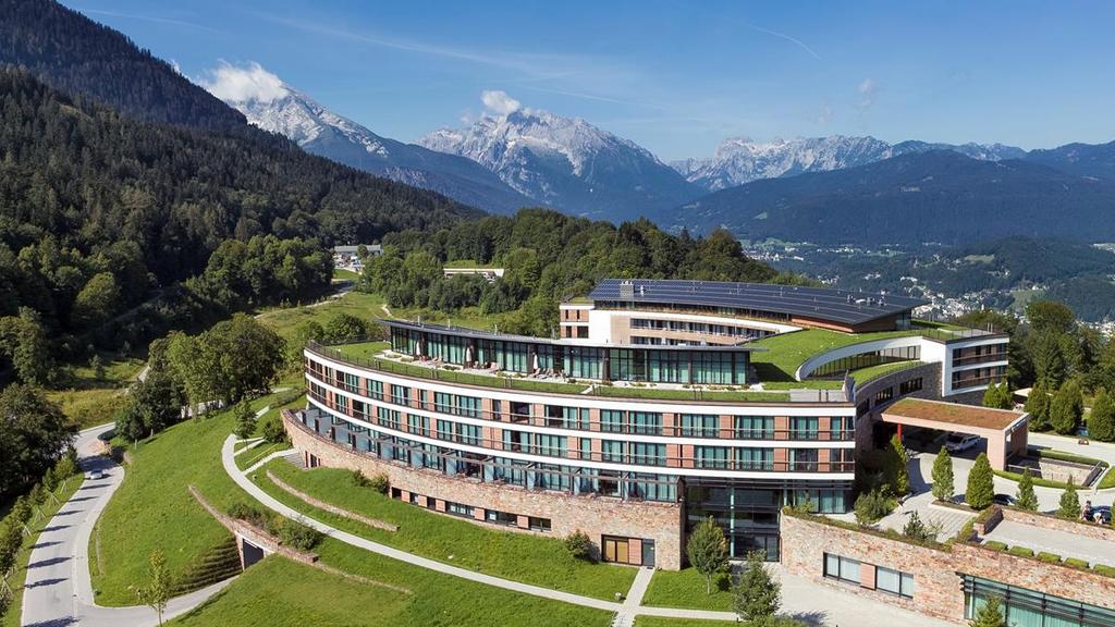 The curved building of Kempinski Berchtesgaden with its green roof among the mountains in summer