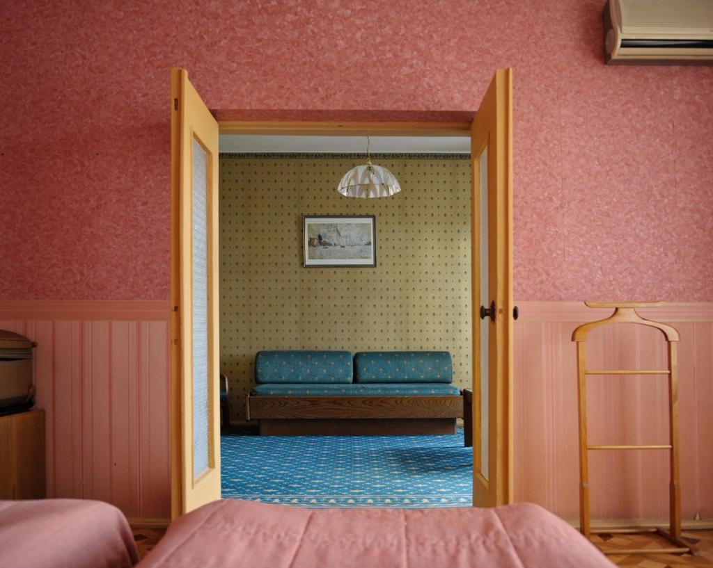 A hotel room interior: pink walls, an open door leading to a room with a blue sofa and a patterned carpet