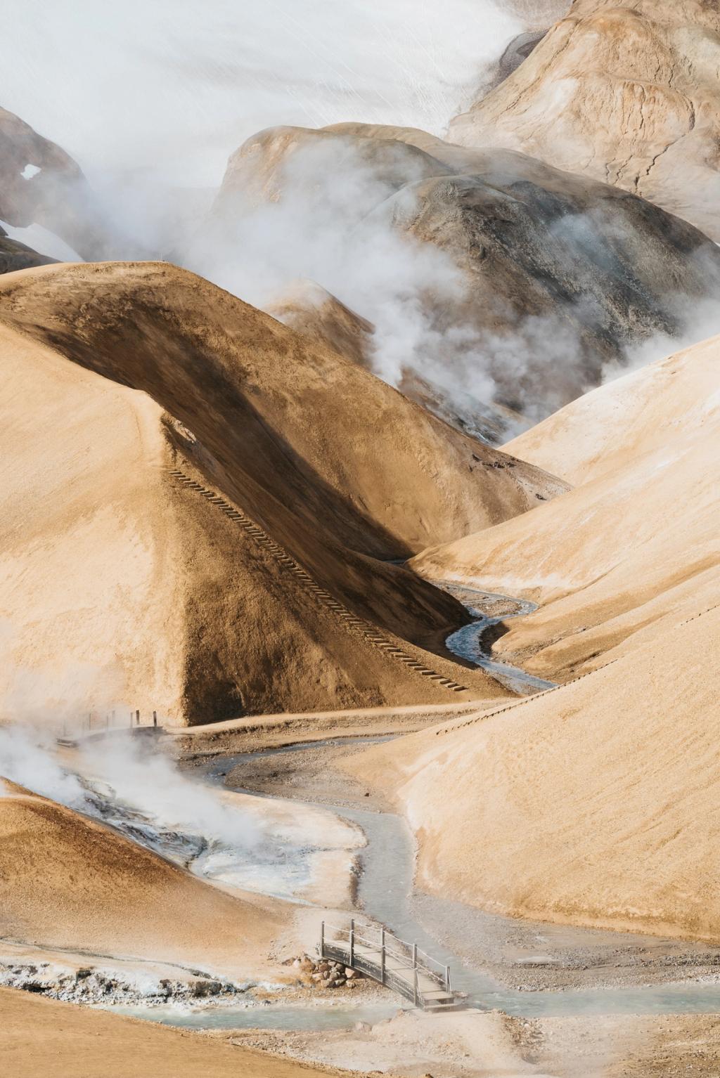 A wooden footbridge over a stream among the sandy hills of Kerlingarfjöll, clouds of steam drifting above a geothermal slope