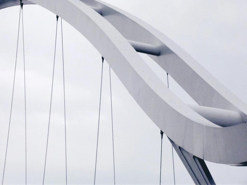 A curved white span of the Infinity Bridge with stay cables against an overcast sky