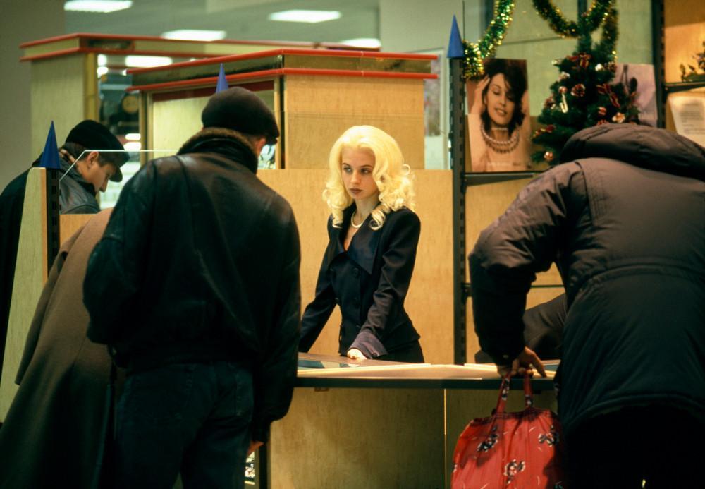 A fair-haired girl behind a counter between customers in winter clothing