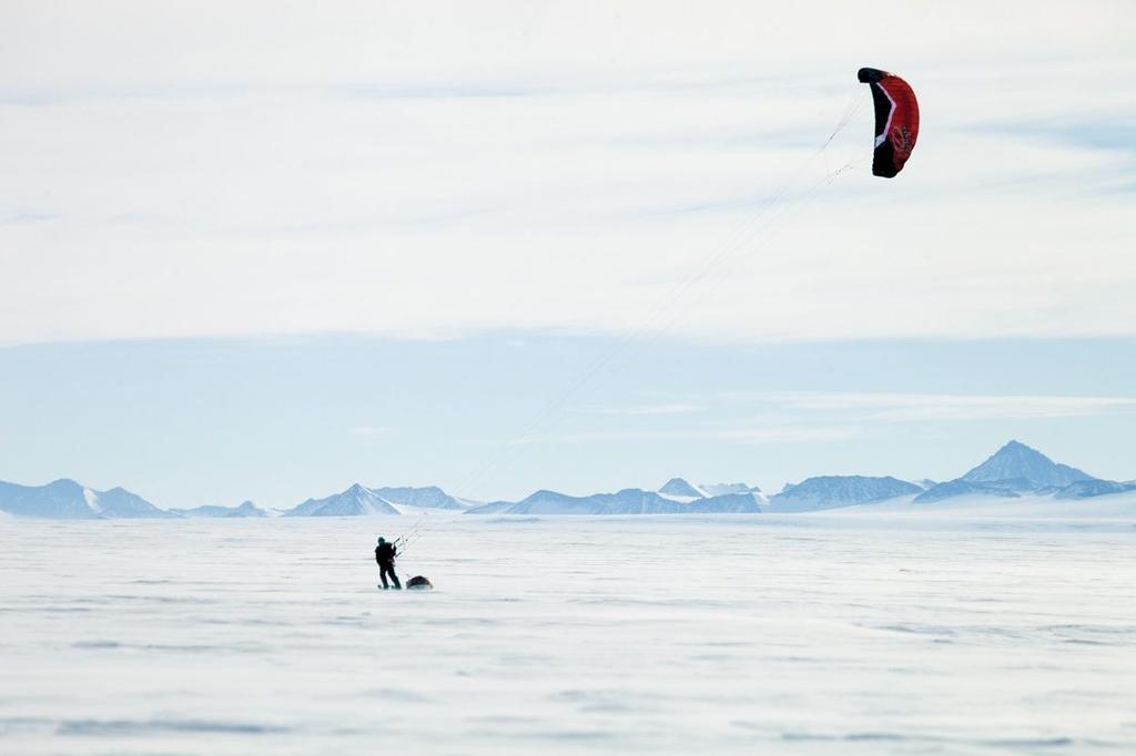 A kitesurfer with a red parafoil gliding across a snowy plain near the mountains