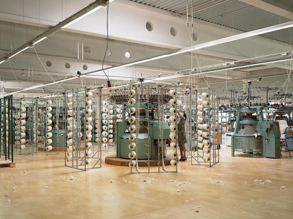 The workshop floor of a textile factory with rows of knitting machines and bobbins of white yarn, a worker at one of the frames