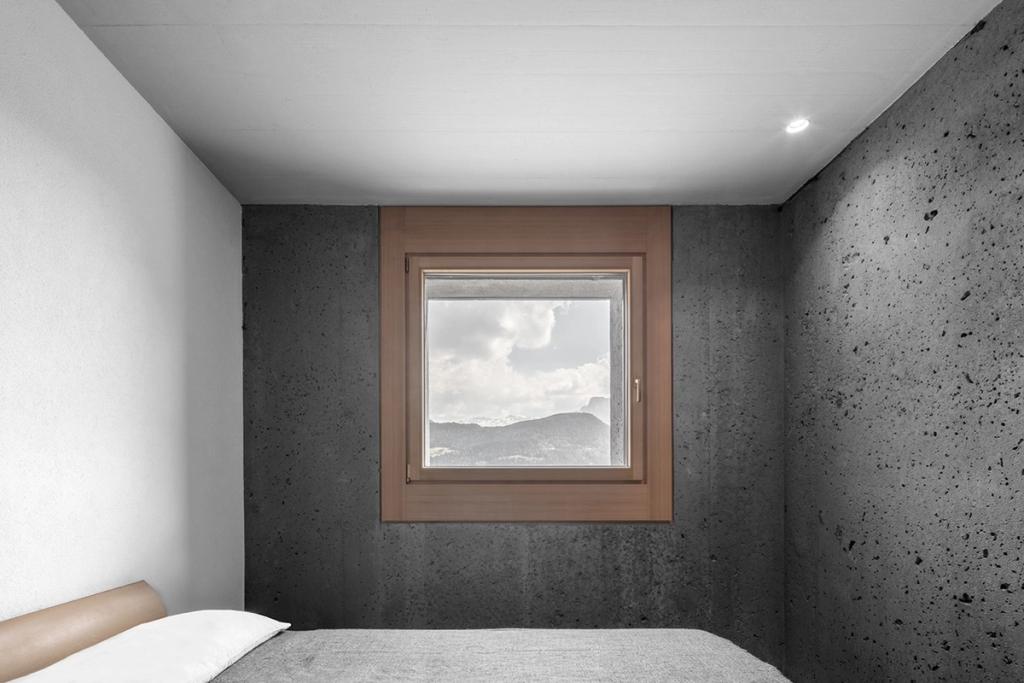 A bedroom with concrete walls and a square window overlooking the mountains