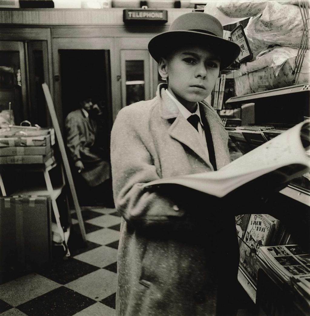 A boy in an adult's hat and coat browsing a magazine at a newsstand