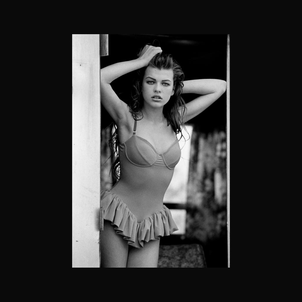 A black-and-white photograph of a young woman in a ruffled swimsuit at a doorway
