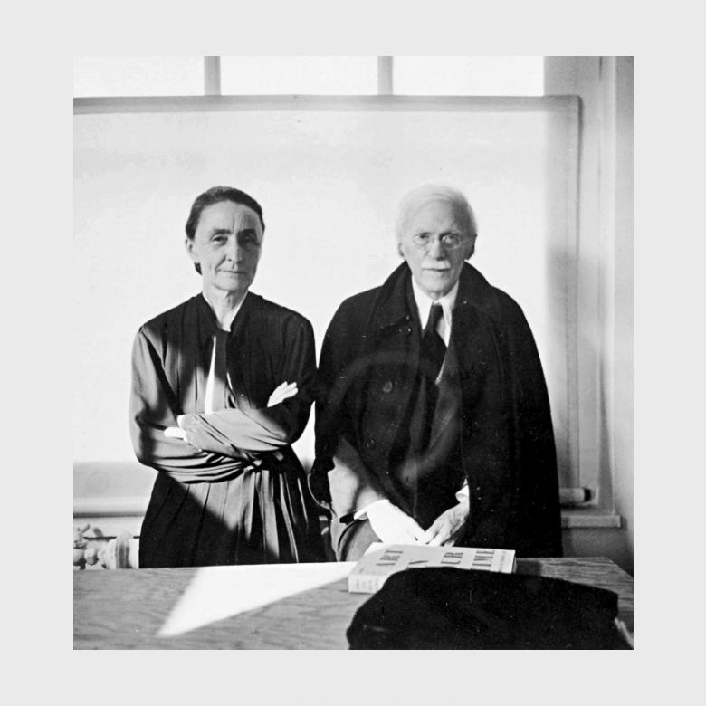 A black-and-white photograph of O'Keeffe with her arms crossed, standing beside Stieglitz