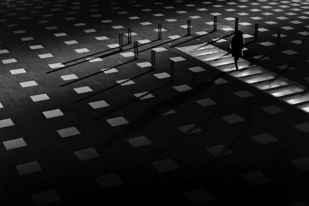 The silhouette of a person climbing steps, the floor scattered with pale squares and the long shadows of posts