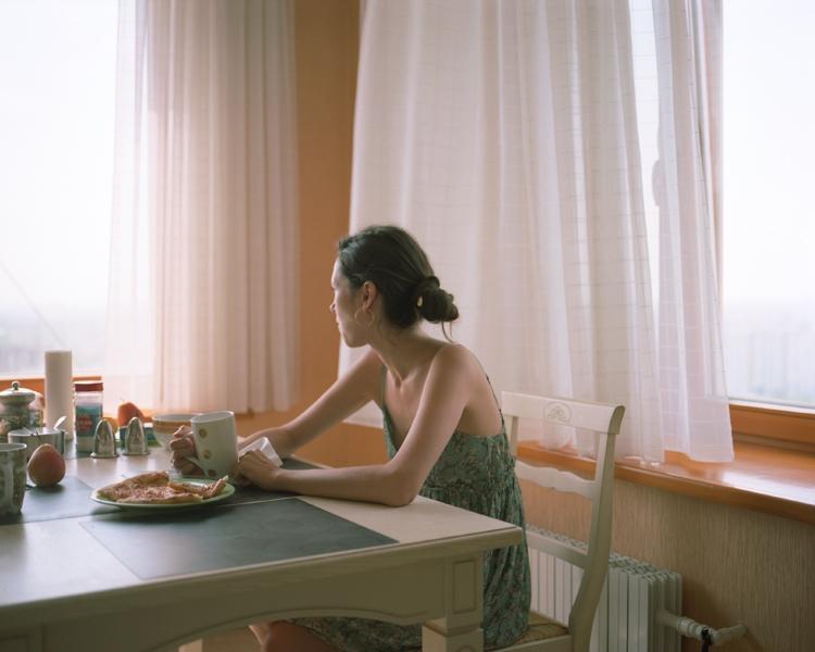 A young woman in a dress sits at breakfast by a window with net curtains in the morning light