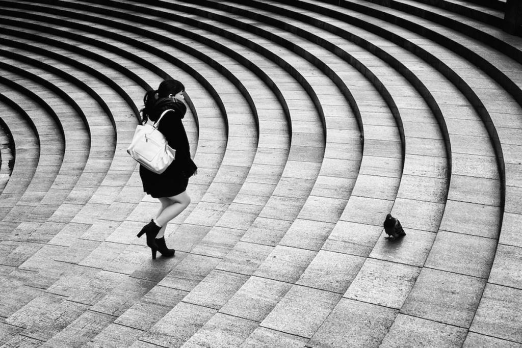 A woman in black, wearing high heels and carrying a white bag, descends semicircular steps, a pigeon nearby