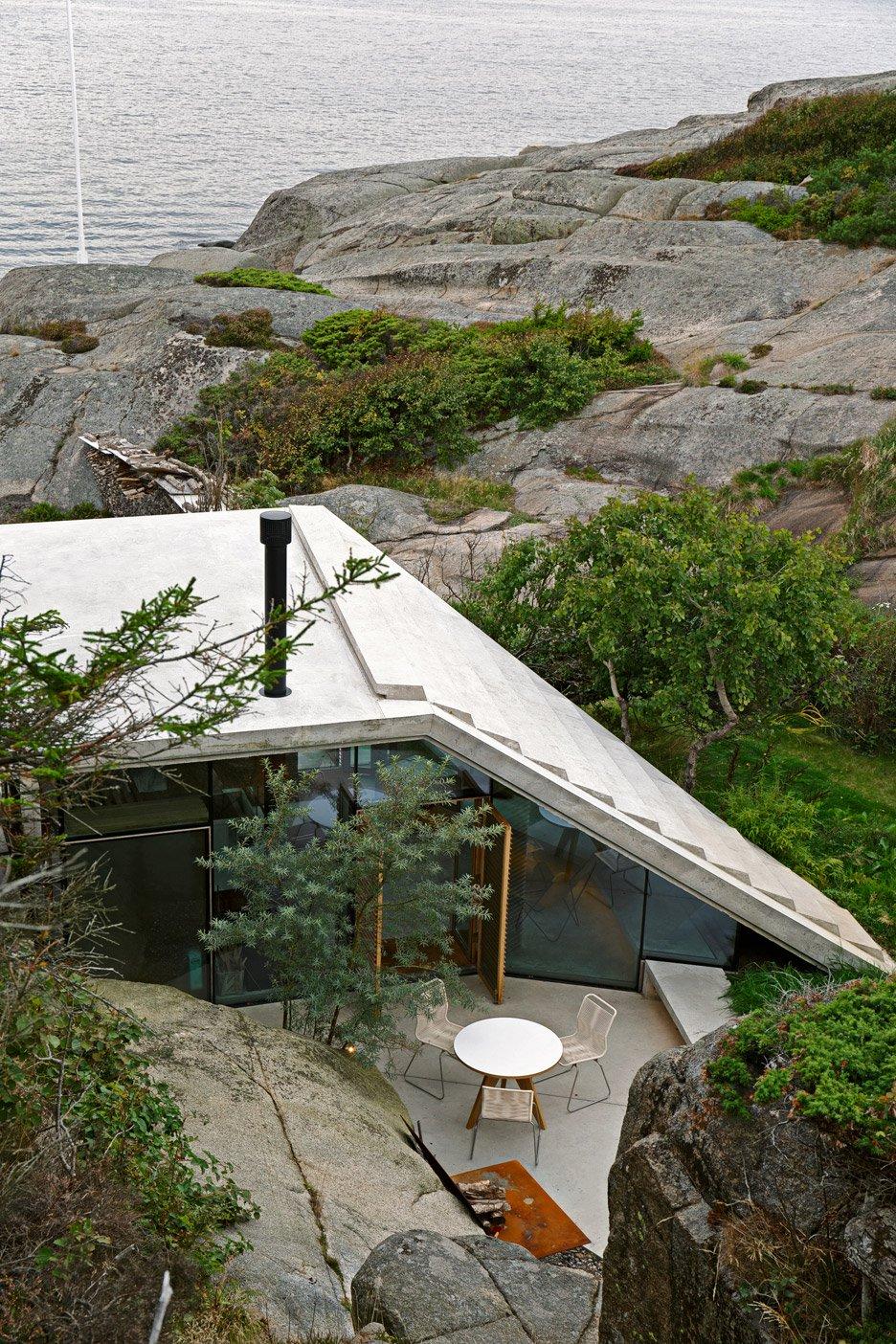 The sloping roof of a glass-walled cabin among granite rocks at the water's edge, aerial view