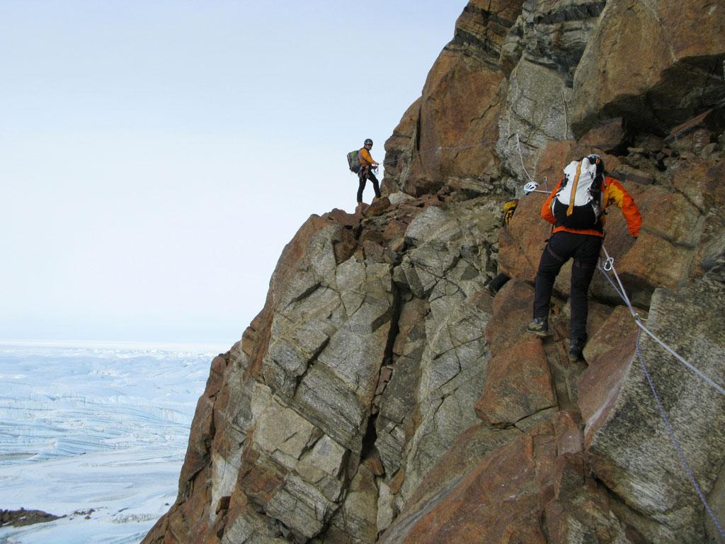 Two climbers ascending a sheer rock face above a glacial plain