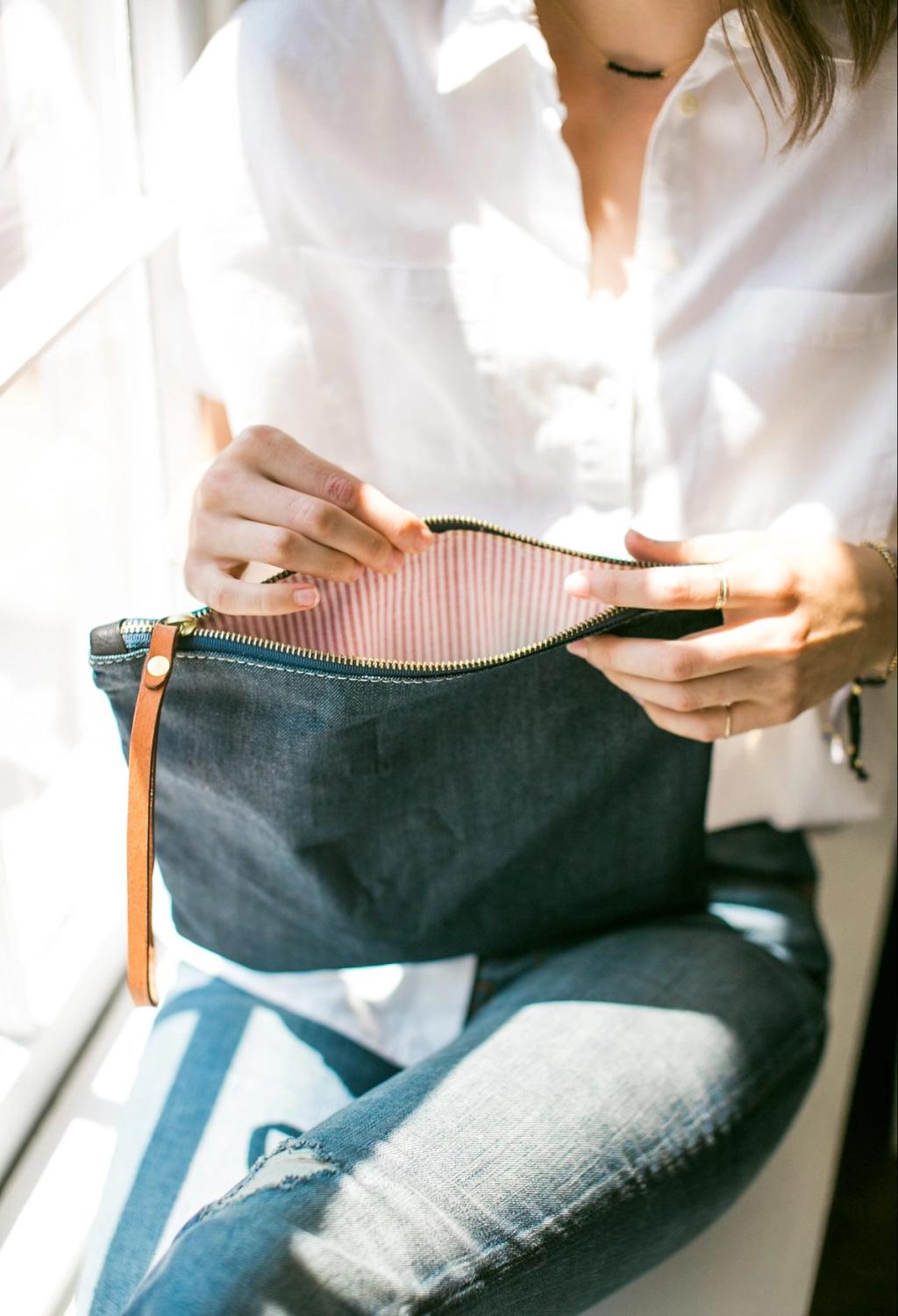 A pair of hands opens a blue denim Tribe Alive clutch bag with a leather strap