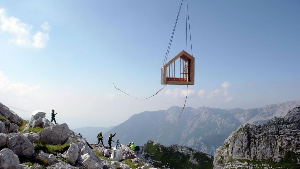 Деревянный модуль бивуака Alpine Shelter Skuta поднимают на вертолётном тросе над скалистым горным хребтом