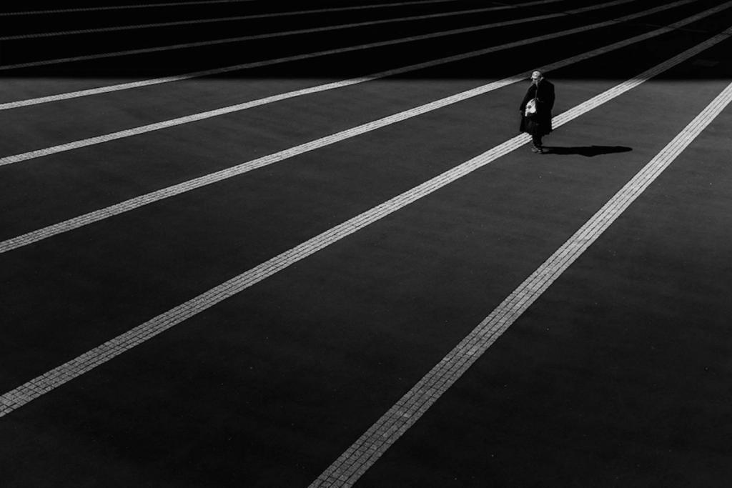 A black-and-white shot of a lone passerby on an empty square, parallel lines of cobblestones receding into the distance