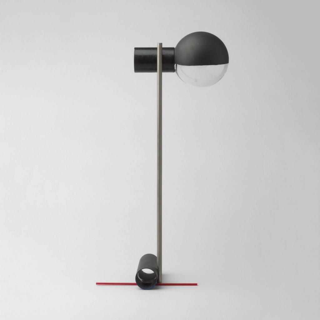A desk lamp with a glass shade on a slender stem and a red counterbalance base