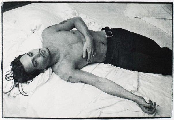 A shirtless man lies on a white bed, a cigarette hanging from his lowered hand