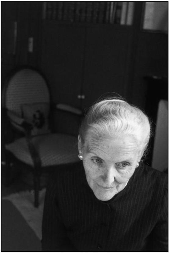 Portrait of a grey-haired elderly woman leaning towards the camera, with an antique armchair beside her