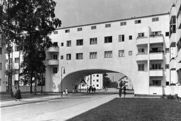 A residential quarter with a drive-through arch beneath a white modernist building; people walking along the street