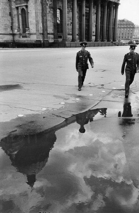 Two Soviet officers walk across a square, their silhouettes reflected in a large puddle