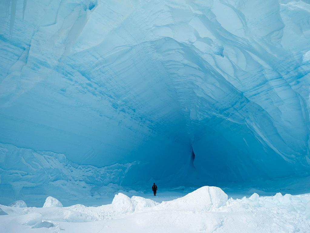 A human figure at the entrance to a vast blue ice cave