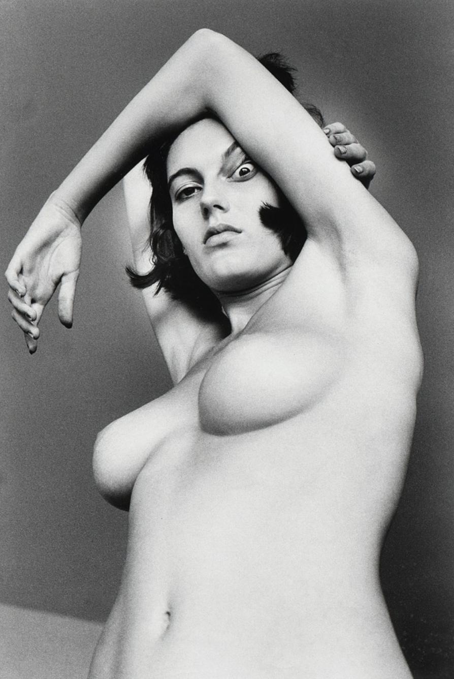 A nude woman with her arms thrown back above her head against a grey wall with a shadow