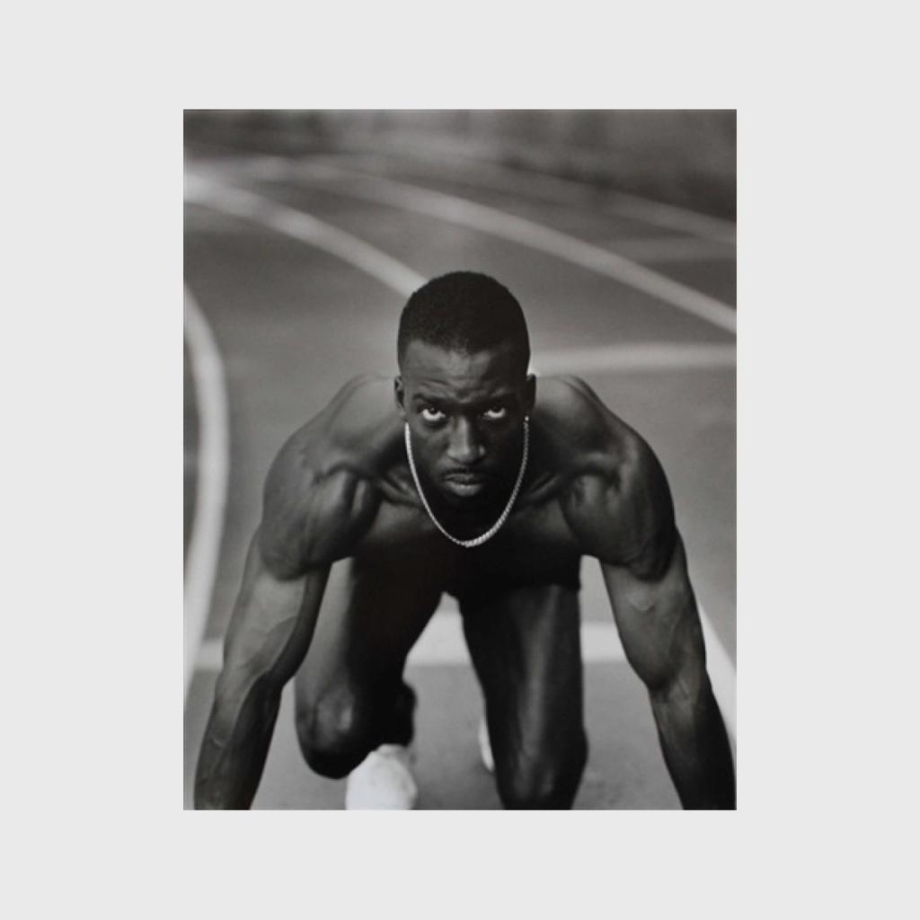 A black-and-white photograph of a muscular sprinter in a starting position on a running track