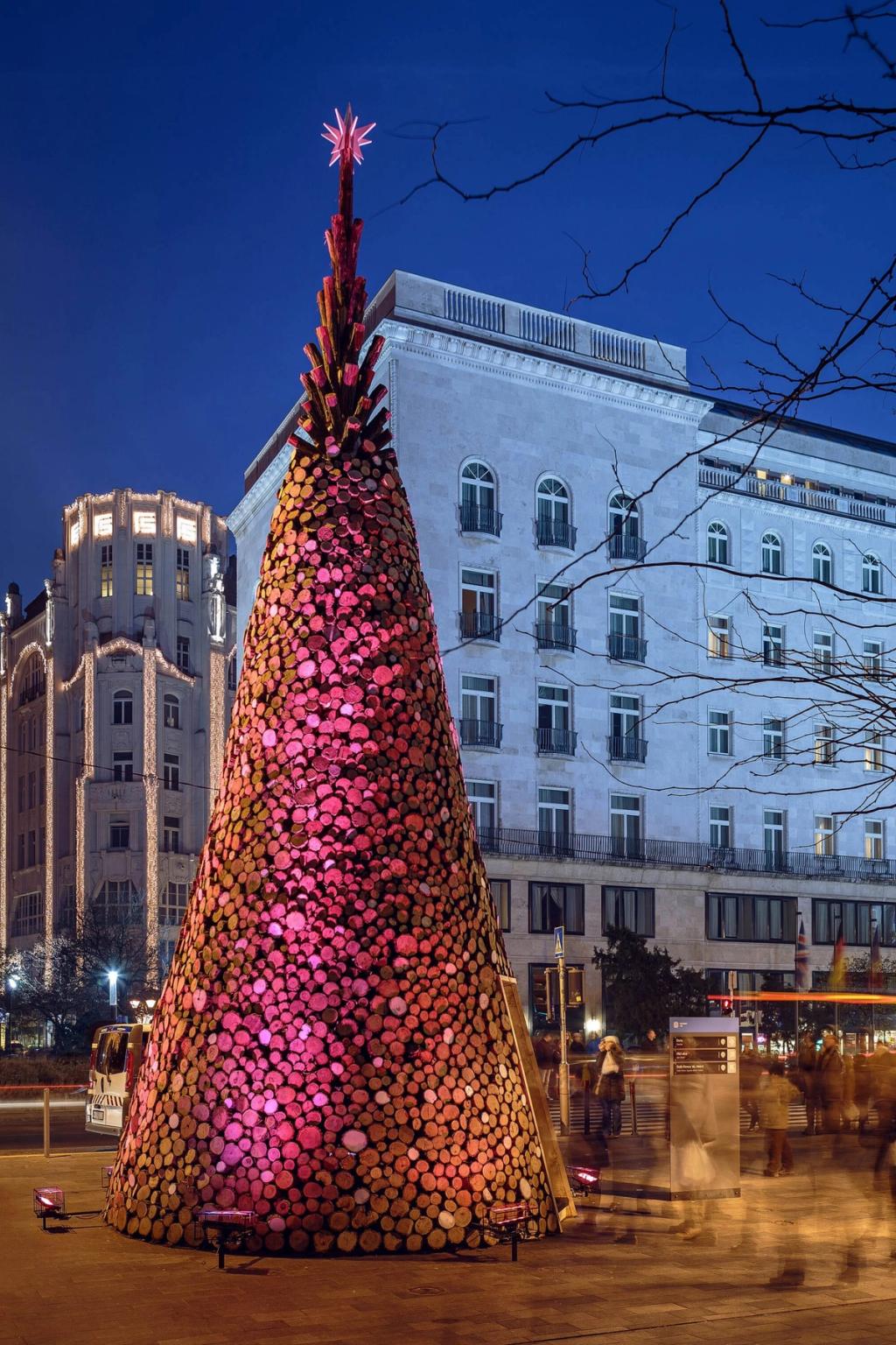 A cone-shaped Christmas tree built from stacked logs, bathed in pink light, on a Budapest square at night