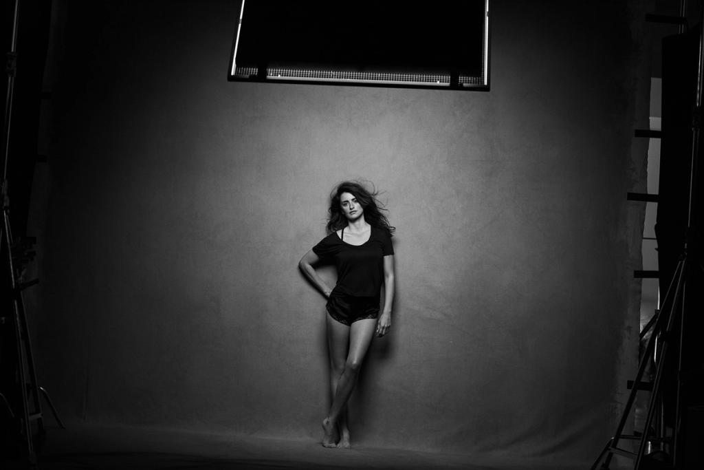 A woman in a black T-shirt and shorts stands barefoot beside a studio wall beneath a softbox