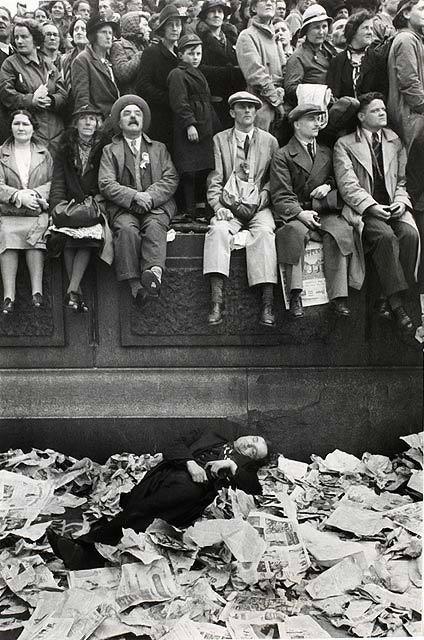 Spectators sit on a plinth; below, a woman lies among scattered newspapers