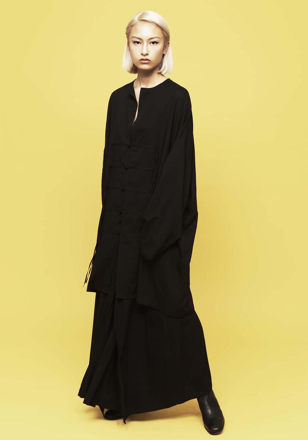 A model in a long black jacket with fastenings and a skirt against a yellow background