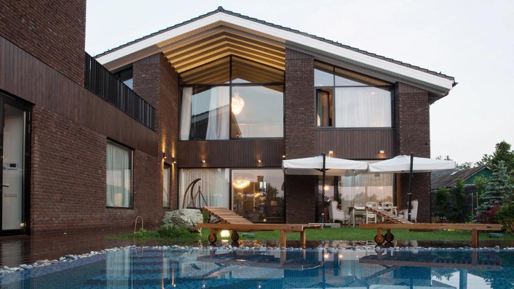 The rear facade of a two-storey brick house with large windows and a pool terrace at dusk