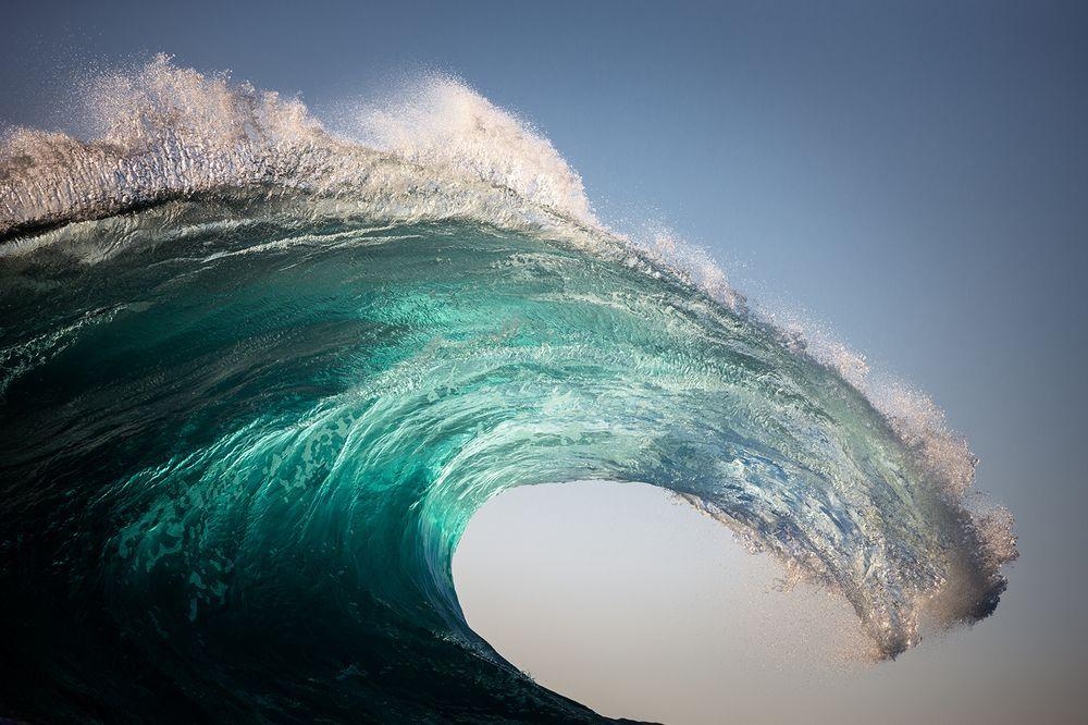 The curving crest of a turquoise ocean wave with spray and foam against a clear sky
