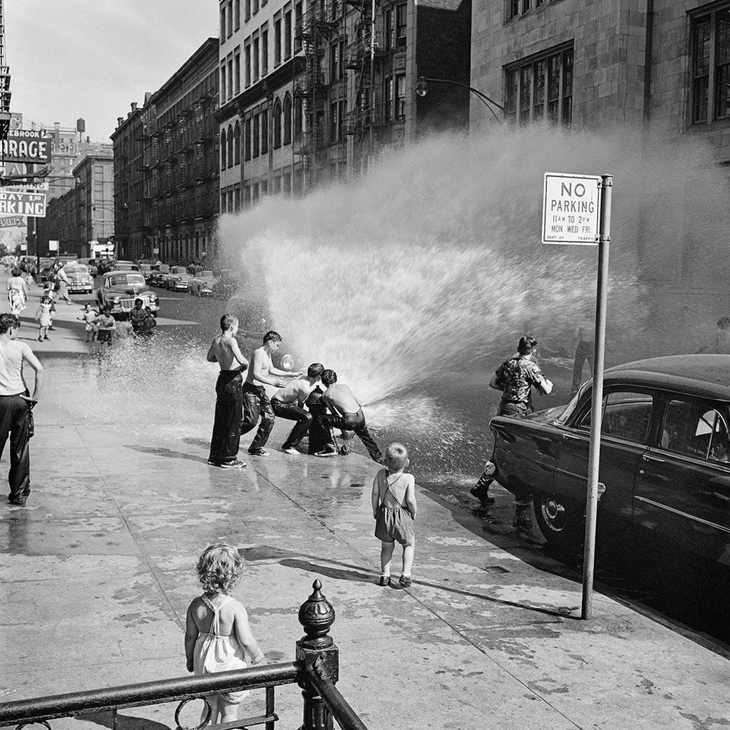 Black-and-white photograph: teenagers and children cool off under a jet of water from a fire hydrant on a summer street