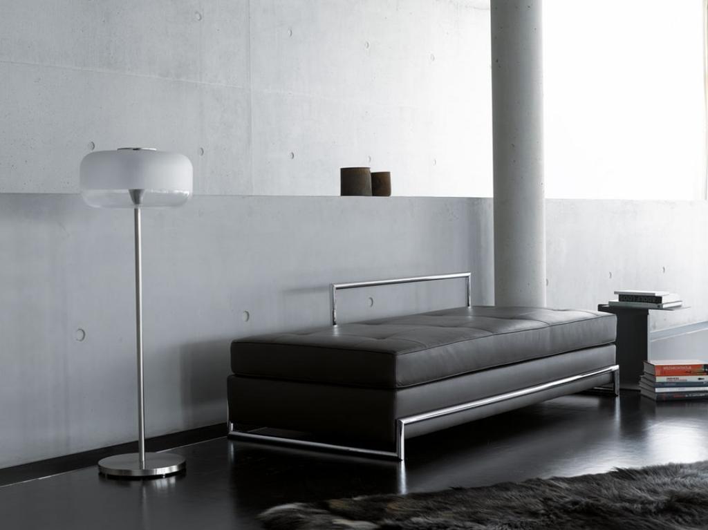 A dark leather daybed on a chromed frame with a floor lamp against a concrete wall