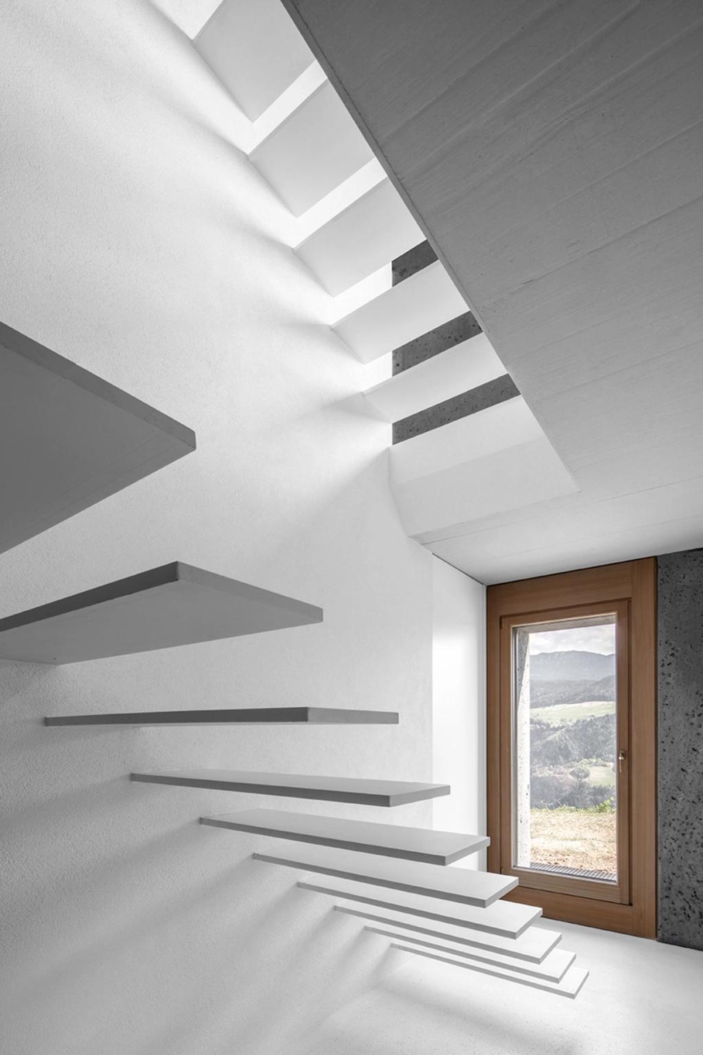 A floating concrete staircase along a white wall and a narrow wooden window with a view of the valley